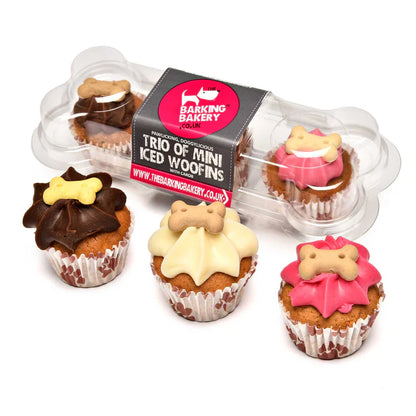 A trio of mini iced woofins from Barking Bakery, each with a different color topping and a dog bone decoration on top, packaged in a clear plastic container.